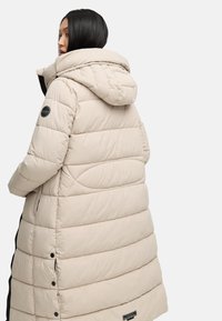 Piumino beige con cappuccio, caratterizzato da un design trapuntato, tasche laterali e un'etichetta con logo sulla manica sinistra. Texture liscia e lunghezza al ginocchio.