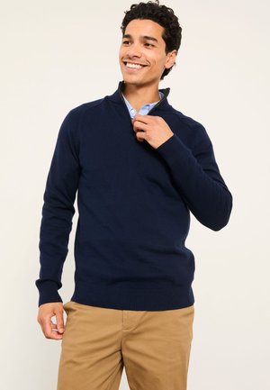 Smiling man in tan pants adjusts the zipper on his navy blue pullover sweater over a light blue shirt, standing against a plain background.
