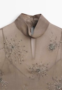 Transparante taupekleurige blouse met hoge kraag, keyhole-halslijn en zilveren kraal bloemversieringen aan de voorkant.