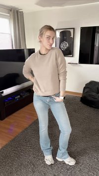 Tânăr care pozează în interior purtând un pulover bej, blugi bleu deschis și adidași albi într-un living cu televizor și opere de artă.