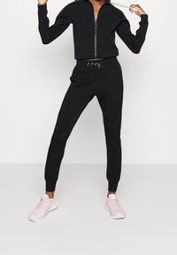 Svart zip-up crop jacket med matchande joggersbyxor, med en slim fit och elastiska ärmslut. Bärs tillsammans med ljusrosa träningsskor.