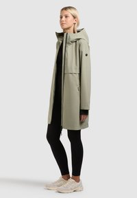 Femme debout portant un imperméable à capuche gris clair sur un legging noir et des baskets blanches, devant un fond uni gris clair.