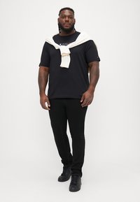 Μαύρο μπλουζάκι με λογότυπο, άσπρη ζακέτα δεμένη γύρω από τους ώμους, μαύρο παντελόνι και μαύρα αθλητικά. Απλός σχεδιασμός, χαλαρό στυλ.