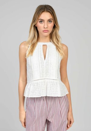 Femme blonde portant un chemisier blanc sans manches en œillet avec un décolleté en trou de serrure et un bas péplum, associé à un pantalon taille haute à rayures rouges et blanches.