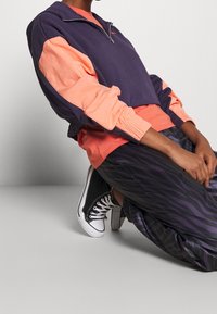 Fialová a korálová mikina na zip s žebrovanými manžetami, doplněná černými a fialovými pruhovanými kalhotami a černými teniskami. Neformální sportovní oblečení.