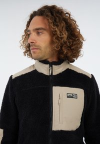 Hombre con cabello rizado que lleva una chaqueta de forro polar negra y beige con bolsillo en el pecho y el logo "PROTEST", mirando hacia la izquierda contra un fondo blanco.