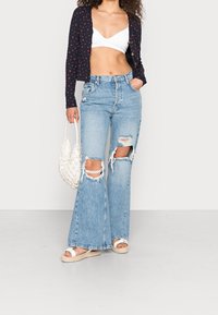 Leichter navyfarbener Cardigan mit kleinen roten Punkten über einem weißen Bralette. Hochtaillierte, ausgewaschene hellblaue Jeans und weiße Sandalen. Geflochtene Tasche.