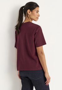 Vrouw met donker haar in een paardenstaart, gekleed in een effen bordeauxrode shirt met korte mouwen en donkere spijkerbroek, staat zijwaarts tegen een witte muur.