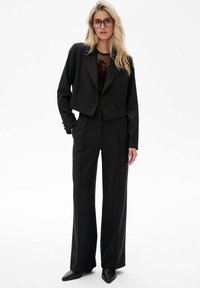 Czarna skórzana marynarka o kroju krótkim z podwójnymi guzikami, zestawiona z czarnymi spodniami o wysokim stanie. Model nosi okulary i czarne buty na szpilkach.