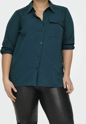 Person wearing dark teal long-sleeve button-up shirt with pocket flap and rolled sleeves, paired with black leather pants, standing.
