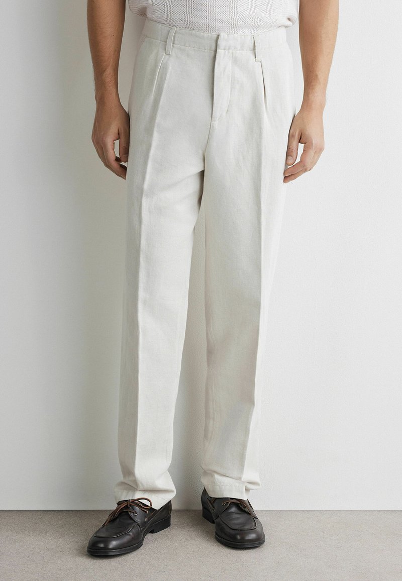 Homme portant un pantalon blanc plissé taille haute et des chaussures en cuir noires à lacets, debout contre un mur blanc uni et un sol gris.
