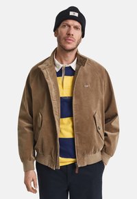 Homem vestindo um gorro preto, uma jaqueta de veludo cotelê castanha sobre uma camisa de rugby listrada em amarelo e azul-marinho, e calças escuras, de pé com as mãos nos bolsos.