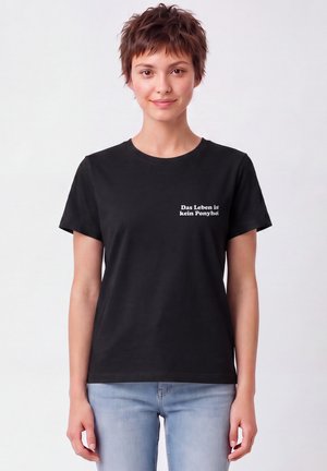 Jeune personne aux cheveux courts bruns portant un t-shirt noir et un jean bleu clair, souriant avec un texte en allemand sur la poitrine gauche.