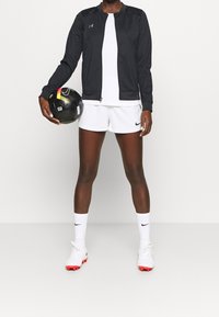 Black athletic jacket over a white shirt, paired with white shorts and socks. Holding a black and yellow soccer ball. White cleats with red accents.