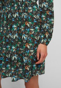 Vestido floral de tela marrón oscuro con patrones de hojas azules, verdes y naranjas; mangas largas y cintura fruncida. Texture suave, a la altura de la rodilla.