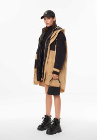 Veste matelassée beige et noire avec capuche, col roulé noir, jupe beige, sac à main noir et bottes à plateforme noires épaisses.
