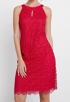 Femme portant une robe rouge en dentelle sans manches avec un décolleté en trou de serrure et un ourlet festonné, debout sur un fond blanc uni.