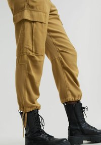 Pantaloni cargo in tessuto beige con multiple tasche, polsini elastici, abbinati a stivaletti neri da combattimento con lacci e cerniera laterale.