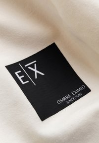 Zwarte label op crème stof met witte tekst: "E|X OMBRE EXIMIO SINCE 1985." Glad textuur met een rechthoekige vorm.