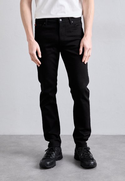 Černé džíny slim fit s klasickým pětipocket designem a kovovými doplňky, v kombinaci s černými sportovními botami.