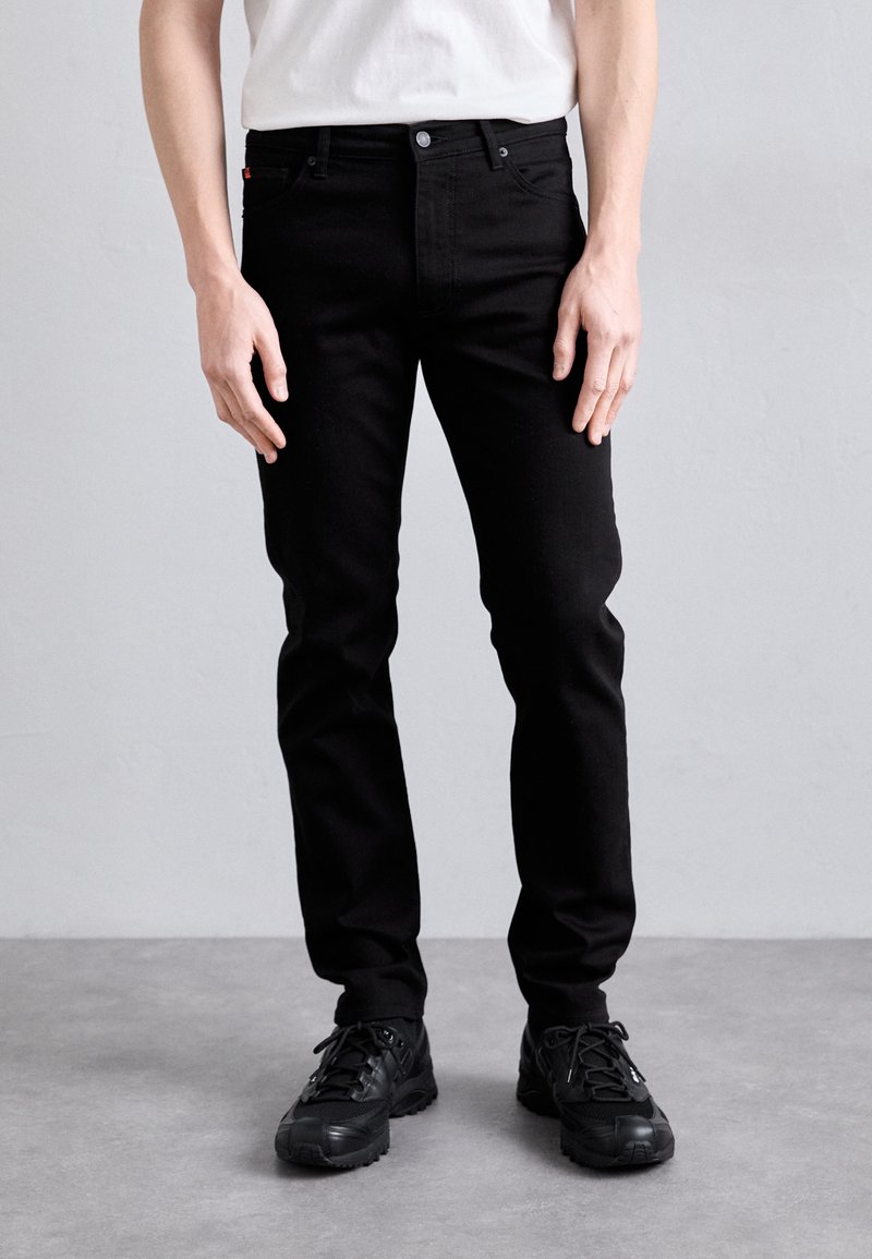 Schwarze Denim-Jeans mit schlanker Passform, ausgestattet mit einem klassischen Fünf-Taschen-Design und Metallbeschlägen, kombiniert mit schwarzen Sport Schuhen.