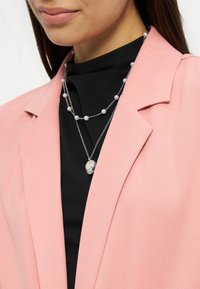 Femme portant des colliers argentés superposés avec des perles et un pendentif, sur un haut noir et un blazer rose.