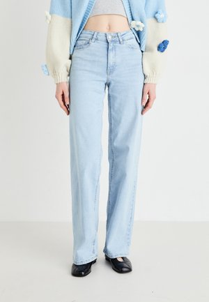 Person trägt hellblaue, hochtaillierte gerade Jeans, kurz geschnittenen hellblauen und weißen Pullover mit floralen Details sowie schwarze Schuhe mit quadratischer Spitze.