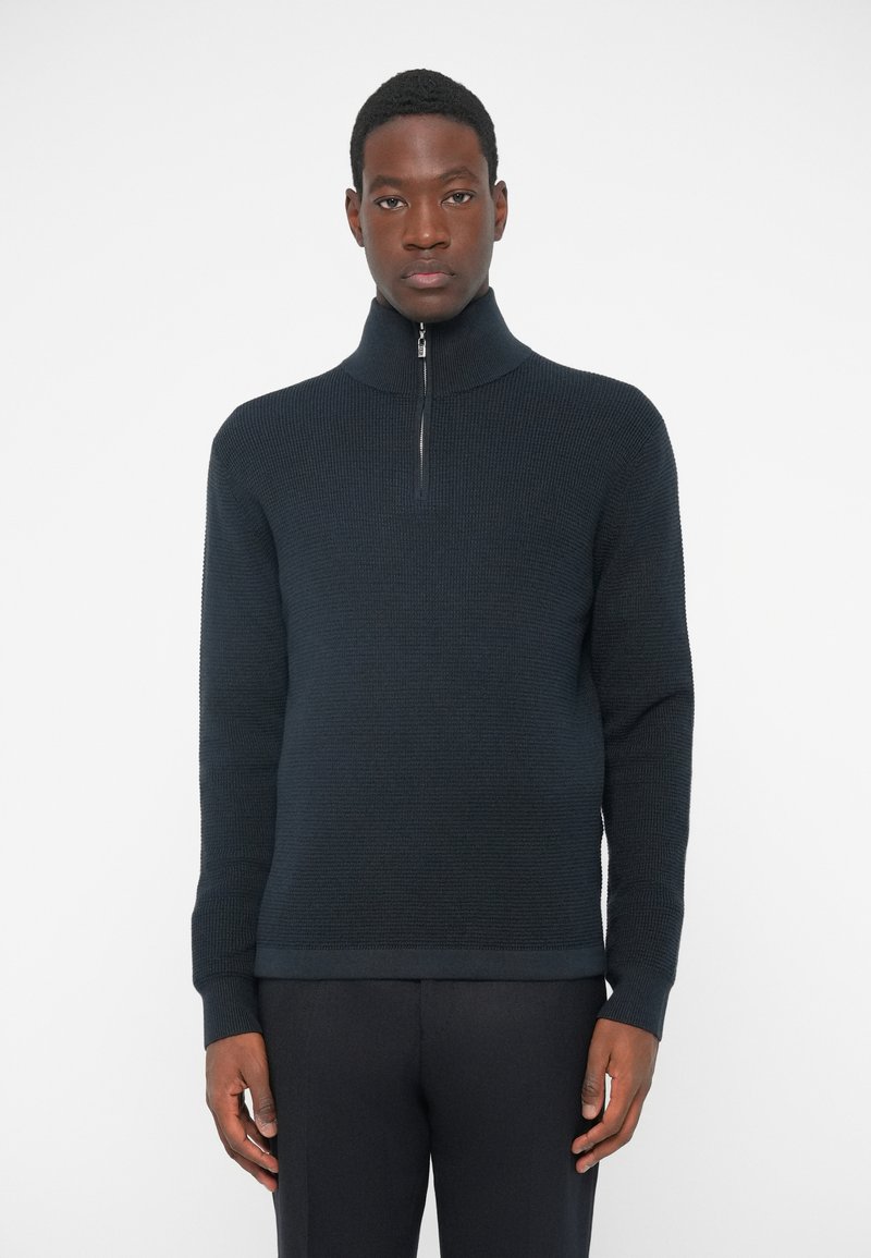 Pulover cu fermoar de culoare bleumarin, cu guler înalt; material texturat, cu nervuri; manșete ajustate; și un design clasic, elegant.