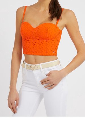 Vrouw draagt een feloranje crop top met eyelets en dunne bandjes, gecombineerd met witte hoog-tailles broek en een beige riem met een driehoekige gesp.