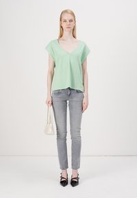 Lysere grønn V-hals topp med korte ermer, kombinert med grå skinny jeans. Modellen holder en liten hvit håndbag og har på seg svarte spisse sko.