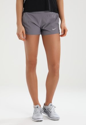 Jambes d'une personne portant un short de sport gris Nike, un haut noir et des chaussures de course blanches, debout sur un fond blanc uni.