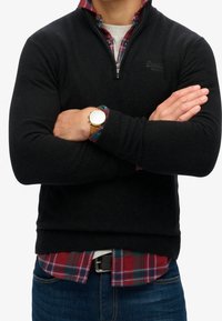 Maglione nero a coste con colletto a mezza zip, indossato sopra una camicia a quadri rossi e verdi. Jeans denim blu scuro con una cintura nera.