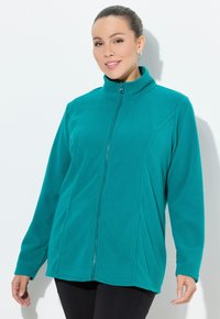 Donna sorridente, che indossa una giacca in pile verde acqua con zip e maniche lunghe e pantaloni neri, in piedi contro uno sfondo bianco.