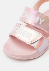 Vaaleanpunaiset liukuvaatereet, joissa on kaksi helmiäishenkistä nauhaa, joissa on valkoiset tekstit "TOMMY HILFIGER." Raskas pohja ja sileä päällismateriaali.