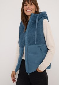 Blaues ärmelloses Vest mit einer flauschigen, strukturierten Oberseite, einem glatten unteren Abschnitt und einer Kapuze. Verfügt über einen Reißverschluss und Taschen.