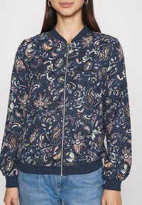 Mörkblå bombarjacka med en färgglad blommig och paisleymönster, dragkedjestängning, ribbade ärmslut och nederkant. Mjuk, texturerad tyg.