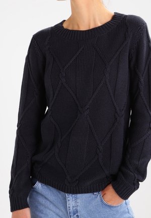 Femme portant un pull en maille torsadée noir avec des motifs en losange et un jean bleu clair, main dans la poche, sur un fond uni.