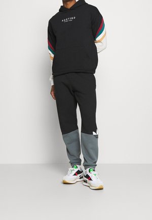 Homme portant un sweat à capuche noir avec des rayures colorées sur les manches et un pantalon de jogging noir et gris, se tenant sur des baskets blanches sur un fond uni.