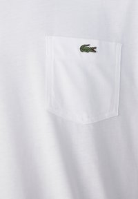 T-shirt en coton blanc avec une poche ornée d'un petit logo de crocodile brodé en vert. Texture lisse avec un col rond classique.