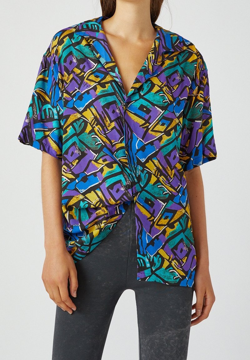 Camisa de manga corta con un patrón abstracto vibrante que presenta colores púrpura, azul, verde y amarillo sobre un fondo negro; corte holgado; tejido ligero.
