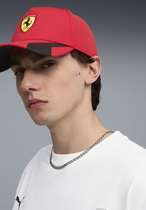Jonge man met een rode Ferrari pet, wit Puma shirt en zilveren kettinkje tegen een effen grijze achtergrond.