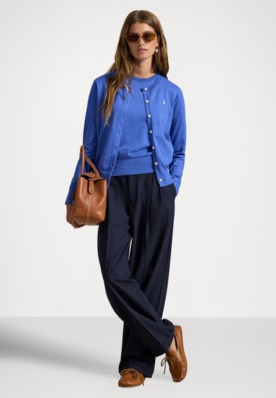 Μπλε ζακέτα και αντίστοιχο πουλόβερ φορεμένα πάνω από σκούρο μπλε παντελόνι. Καφέ δερμάτινη τσάντα με καμπυλωτό σχεδιασμό. Το μοντέλο φορεί γυαλιά ηλίου και casual παπούτσια.