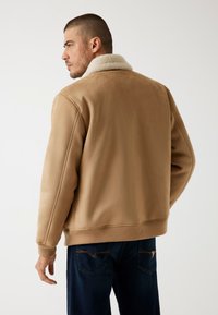 Beige Fleece-Jacke mit weicher Textur, Rippbündchen und -saum sowie klassischem Kragen, aus der Rückansicht präsentiert. Dunkle Jeans sind darunter sichtbar.