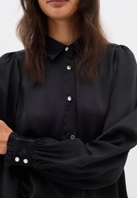 Musta satiiniblouse, jossa on kauluksellinen pääntie, nappilista, jossa on kristallimaiset napit, ja tilavat puffihihat hihansuissa.