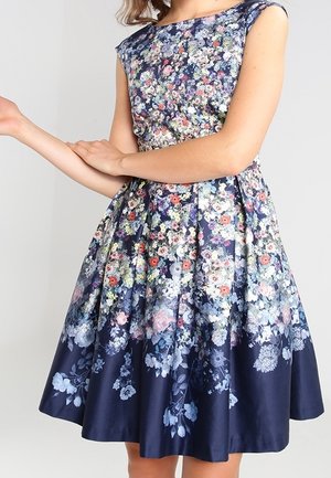 Vrouw draagt mouwloze marineblauwe jurk met meerkleurig bloemenpatroon en aansluitend lijfje, handen gekruist in de taille.