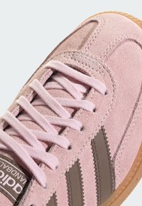 Sapatilha de camurça rosa com atacadores rosa claro, riscas laterais em couro castanho e sola de borracha bege. Apresenta detalhes cosidos e um acabamento texturizado.