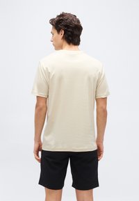 Νεαρός άνδρας με σκούρα κυματιστά μαλλιά, που στέκεται γυρισμένος προς τα πίσω, φορώντας μια απλή μπεζ κοντομάνικη μπλούζα και μαύρο σορτς, μπροστά σε λευκό φόντο.