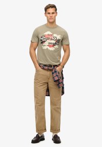 Olijfgroene T-shirt met 'Superdry' grafiek, gecombineerd met tan chino's en een rood-blauw geruit overhemd om de taille gebonden. Donkere loafers.