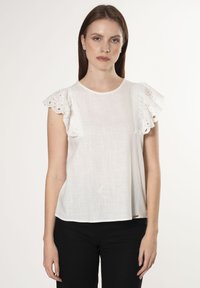 Blusa bianca con maniche a sbuffo in pizzo ad occhio di pernice, scollatura rotonda e tessuto morbido e testurizzato. Indossata con pantaloni neri. Sfondo semplice.