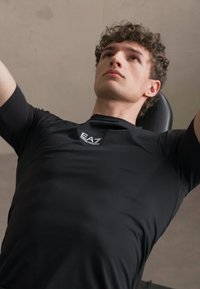 Svart treningstrøye med tettsittende design, glatt tekstur, og logo på brystet. Person trener på en benk for vekter.